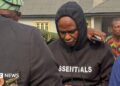 Adeniyi Mobolaji Kayode is in a black hoody with his head bowed as someone has their arm round him as he is led from court in a picture taken in January 20 this year