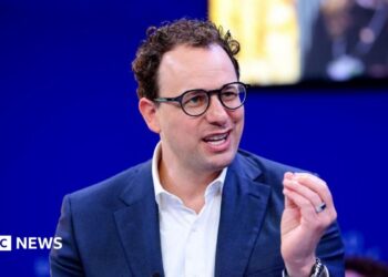 Dario Amodei hld the closed fingers of his left hand together as he mades a point. He is wearing a white shirt and blue jacket, black-rimmed glasses and has slightly curly hair. He is sat against a blue background, with a yellow-lit square just behind and to the left of his head.