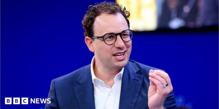 Dario Amodei hld the closed fingers of his left hand together as he mades a point. He is wearing a white shirt and blue jacket, black-rimmed glasses and has slightly curly hair. He is sat against a blue background, with a yellow-lit square just behind and to the left of his head.