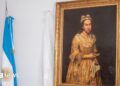 A portrait hangs on the wall with a gold frame on a white backdrop, next to it is an Argentinian flag rolled up
