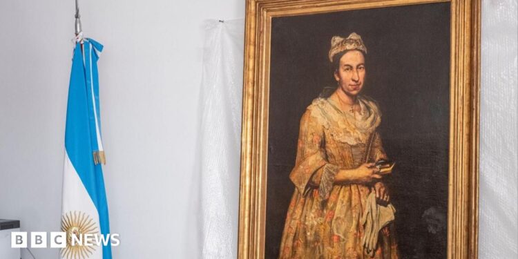 A portrait hangs on the wall with a gold frame on a white backdrop, next to it is an Argentinian flag rolled up