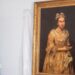 A portrait hangs on the wall with a gold frame on a white backdrop, next to it is an Argentinian flag rolled up