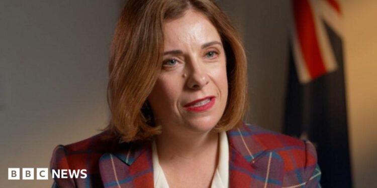 A woman in a tartan jacket and white dress speaks. Several flags can be seen in the background