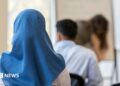 A stock image of a girl wearing a blue headscarf. She is sitting down in a classroom, facing with her back towards the camera.