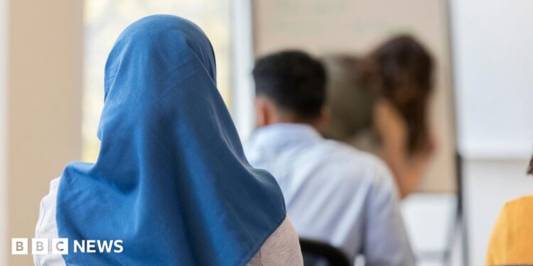 A stock image of a girl wearing a blue headscarf. She is sitting down in a classroom, facing with her back towards the camera.