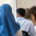 A stock image of a girl wearing a blue headscarf. She is sitting down in a classroom, facing with her back towards the camera.