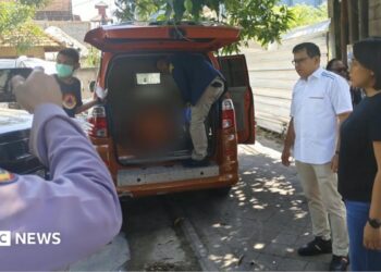 A person stands inside the rear end of an orange van while several other people stand beside the vehicle