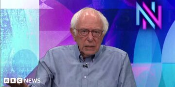 US Senator Bernie Sanders wears a blue button up and sits in front of a green screen projecting a blue and purple graphic and the 'NN' Newsnight logo