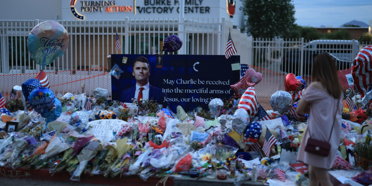 A woman in a pink dress approaches a makeshift memorial for Charlie Kirk outside a Turning Point USA office in Arizona at dusk
