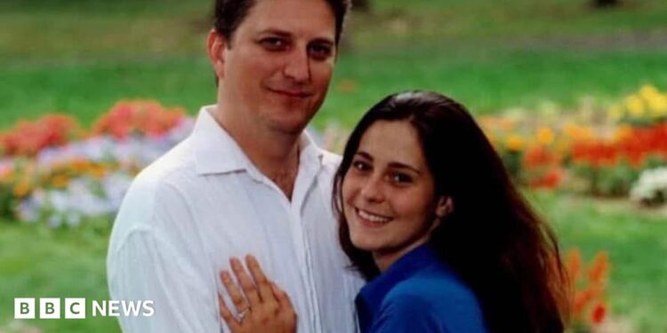 John Resta is on the left, he has dark hair and is wearing a white linen shirt and his wife Sylvia is on the right, with dark long hair and a blue short-sleeved shirt. She is pressed against him, her left hand on his chest. It has a wedding ring on it. They are both smiling. Behind them is grass with flowers.