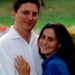 John Resta is on the left, he has dark hair and is wearing a white linen shirt and his wife Sylvia is on the right, with dark long hair and a blue short-sleeved shirt. She is pressed against him, her left hand on his chest. It has a wedding ring on it. They are both smiling. Behind them is grass with flowers.