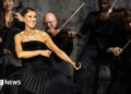 Dion dancing in a black dress, holding a microphone, while musicians play behind her