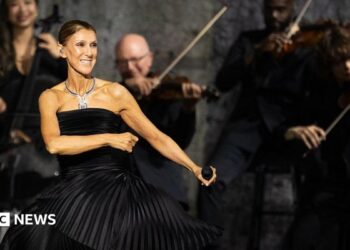 Dion dancing in a black dress, holding a microphone, while musicians play behind her
