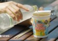 Woman's hand pouring water from a bottle into a cup noddle sitting an a wooden table outside