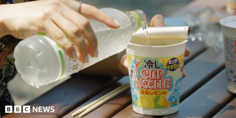 Woman's hand pouring water from a bottle into a cup noddle sitting an a wooden table outside