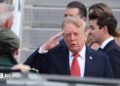 Donald Trump saluting. He is wearing a dark suit with a red striped tie.