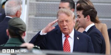 Donald Trump saluting. He is wearing a dark suit with a red striped tie.