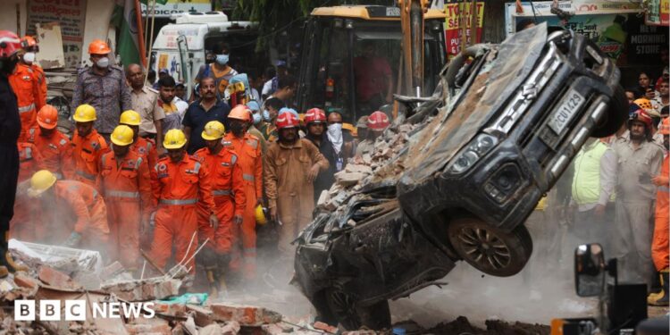 National Disaster Response Force (NDRF) personnel look on at a building collapse in Delhi, while a crushed car is pulled by a digger from the rubble.