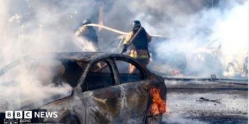 Two firefighters direct a hose at billowing smoke in front of them. A burned-out car is on fire in the foreground.
