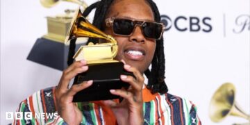 Lil Durk wearing sunglasses and a multi-coloured suit, and holding up the gold gramophone-shaped Grammy Award
