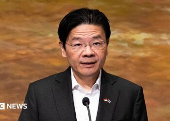 Lawrence Wong in a black suit and white shirt addresses a crowd on a podium