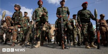 A dozen Houthi men wearing combat clothes, berets and holding large guns. The sky is blue and the men are looking straight ahead.