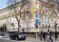 A building in the centre of Budapest surrounded with a metal fence. Five people stand on the pavement at the corner of a road where three cars are seen parked.
