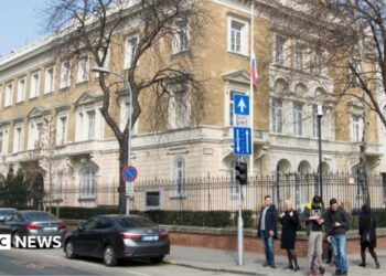 A building in the centre of Budapest surrounded with a metal fence. Five people stand on the pavement at the corner of a road where three cars are seen parked.