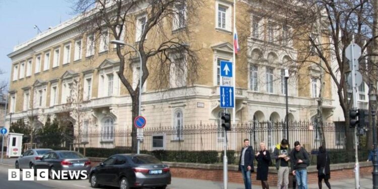 A building in the centre of Budapest surrounded with a metal fence. Five people stand on the pavement at the corner of a road where three cars are seen parked.