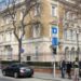 A building in the centre of Budapest surrounded with a metal fence. Five people stand on the pavement at the corner of a road where three cars are seen parked.