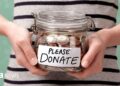 A shot of someone's torso, in a black and white striped top, holding a glass jar with coins in it. On the front is a label that reads: Please donate.