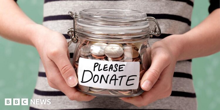 A shot of someone's torso, in a black and white striped top, holding a glass jar with coins in it. On the front is a label that reads: Please donate.