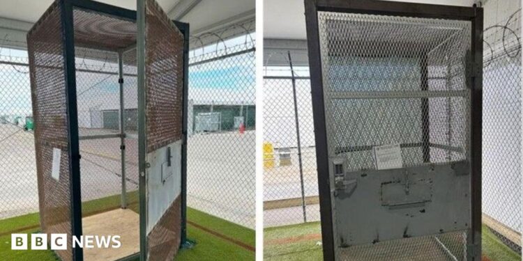A metal cage sits within a larger cage that has razor wire across the top. The small cage has a door with a combination lock
