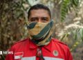 A man in red firefighting uniform patched with Indonesia's flag, and with a yellow and green neck gaiter pulled over his face, stands in a forest and looks into the camera