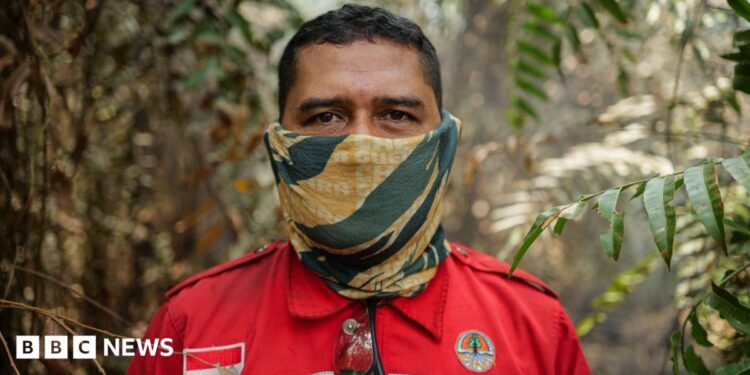 A man in red firefighting uniform patched with Indonesia's flag, and with a yellow and green neck gaiter pulled over his face, stands in a forest and looks into the camera