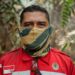 A man in red firefighting uniform patched with Indonesia's flag, and with a yellow and green neck gaiter pulled over his face, stands in a forest and looks into the camera