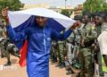 A man in a blue boubou drapes a Russian flag over his head. A group of people in army uniforms can be seen to the right of the picture.