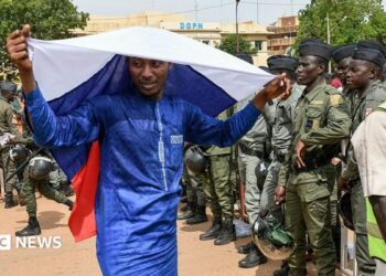 A man in a blue boubou drapes a Russian flag over his head. A group of people in army uniforms can be seen to the right of the picture.