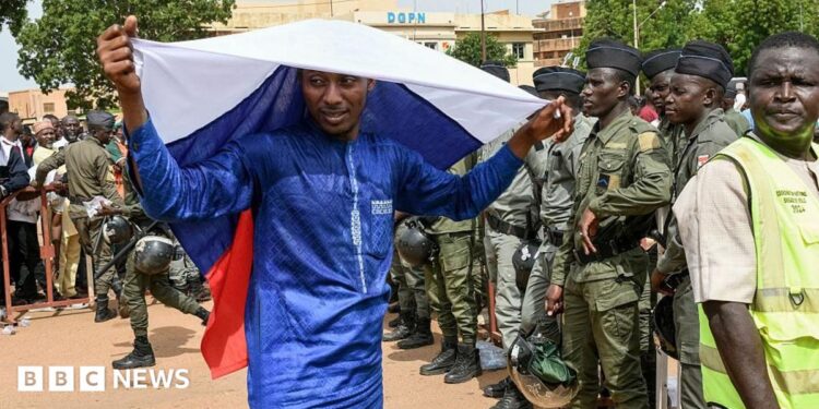A man in a blue boubou drapes a Russian flag over his head. A group of people in army uniforms can be seen to the right of the picture.