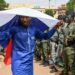 A man in a blue boubou drapes a Russian flag over his head. A group of people in army uniforms can be seen to the right of the picture.
