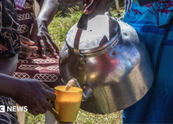 A close-up of someone's hand holding a large stove-top kettle and pouring a brown milky liquid into a yellow cup being held by someone else.