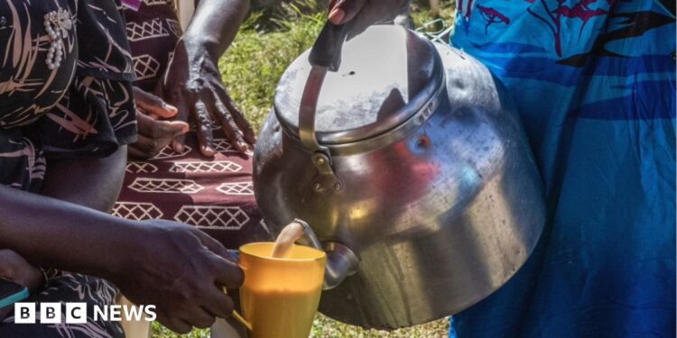 A close-up of someone's hand holding a large stove-top kettle and pouring a brown milky liquid into a yellow cup being held by someone else.