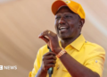 William Ruto in a yellow shirt and cap gestures with his hand as he makes a speech holding a microphone in his other hand.