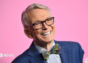 Bob Mackie wears a navy blue blazer, blue and white striped shirt and a multi-coloured tartain style bow-tie. He's smiling and there's a bright pink background.