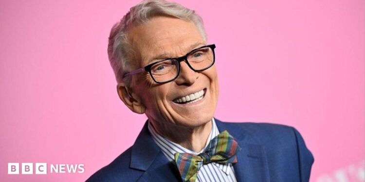 Bob Mackie wears a navy blue blazer, blue and white striped shirt and a multi-coloured tartain style bow-tie. He's smiling and there's a bright pink background.