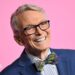 Bob Mackie wears a navy blue blazer, blue and white striped shirt and a multi-coloured tartain style bow-tie. He's smiling and there's a bright pink background.