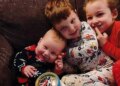 Three young Clancy children in pajamas sit together on a couch, the smiling baby resting against his older brother, with their sister beside them.