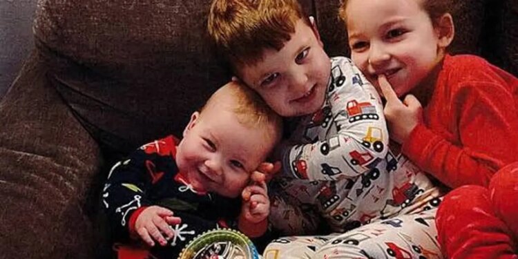 Three young Clancy children in pajamas sit together on a couch, the smiling baby resting against his older brother, with their sister beside them.