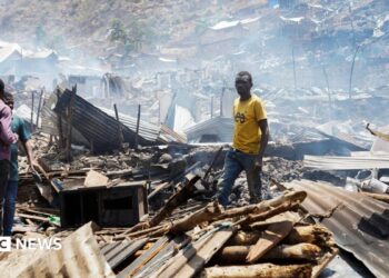 Man in yellow t-shirt stands in piles of rubble and corrugated iron while three other people stand on the left of the picture. The rubble seems to go on for some distance and the air is hazy.