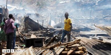 Man in yellow t-shirt stands in piles of rubble and corrugated iron while three other people stand on the left of the picture. The rubble seems to go on for some distance and the air is hazy.
